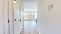 This thoughtfully designed 4-bedroom, 1-bathroom unit spans a single floor and delivers a clean, contemporary aesthetic throughout. Light-toned hardwood flooring flows seamlessly across all rooms, complemented by crisp white walls and modern black hardware accents on doors and fixtures. The open-concept kitchen and living area serves as the heart of the unit, showcasing a large central island with a white quartz countertop and warm wood-grain base, a matte black pull-down faucet, and a full suite of stainless steel appliances including a range, built-in microwave, and refrigerator. Sleek white upper cabinetry and matching wood-toned lower cabinets complete the kitchen, which is illuminated by striking black-and-gold pendant lights. The living area benefits from an expansive bank of large windows that flood the space with natural light and offer open urban views of the surrounding cityscape. Each of the four bedrooms is well-proportioned, featuring the same light hardwood floors and white panel doors with matte black hardware. The shared bathroom is elegantly appointed with full marble-look tile surround, a soaking tub, and a matte black wall-mounted faucet, adding a luxurious touch to the overall design.
