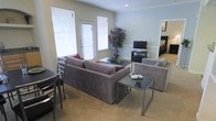 This single-floor space centers around a thoughtfully arranged living area that combines comfort and functionality. The living room features a generously sized sectional sofa in a neutral gray upholstery, complemented by a matching armchair, creating an inviting seating arrangement. Sleek, metal-framed coffee and side tables add a modern touch to the space. The room is finished with wall-to-wall carpeting in a warm beige tone, contributing to the overall cozy atmosphere.

Natural light fills the space through multiple windows fitted with white horizontal blinds, allowing for adjustable brightness throughout the day. The walls are painted in a soft, muted gray-blue tone, providing a calm and neutral backdrop. A decorative tree adds a natural element and visual interest to the corner of the room.

An entertainment setup, including a flat-screen television on a low media console, is positioned along one wall. A glimpse into an adjacent room suggests a bedroom nearby, indicating a well-connected floor plan. The overall ambiance is clean, modern, and livable, making it a practical and welcoming environment.