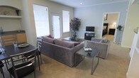 This single-floor space centers around a thoughtfully arranged living area that combines comfort and functionality. The living room features a generously sized sectional sofa in a neutral gray upholstery, complemented by a matching armchair, creating an inviting seating arrangement. Sleek, metal-framed coffee and side tables add a modern touch to the space. The room is finished with wall-to-wall carpeting in a warm beige tone, contributing to the overall cozy atmosphere.

Natural light fills the space through multiple windows fitted with white horizontal blinds, allowing for adjustable brightness throughout the day. The walls are painted in a soft, muted gray-blue tone, providing a calm and neutral backdrop. A decorative tree adds a natural element and visual interest to the corner of the room.

An entertainment setup, including a flat-screen television on a low media console, is positioned along one wall. A glimpse into an adjacent room suggests a bedroom nearby, indicating a well-connected floor plan. The overall ambiance is clean, modern, and livable, making it a practical and welcoming environment.