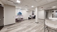 Located at 1225 Santa Fe Drive within a Denver business center, this single-floor office suite offers a clean, modern workspace designed for productivity. The open-plan layout accommodates multiple workstations, each separated by low-profile cubicle dividers and outfitted with sleek white desks and ergonomic task chairs — providing both collaborative energy and a degree of individual focus.

The interior design is defined by a neutral, professional palette: crisp white walls, light gray wood-style plank flooring, and a drop-panel ceiling fitted with flush, evenly spaced recessed LED lighting that ensures consistent, well-distributed illumination throughout the space. A structural column near the center of the room adds an architectural element without disrupting the functional flow.

A high-top table with bar-height seating near the entrance provides a casual meeting or break area. The suite also includes space for a printer and storage unit, supporting day-to-day office operations. Secure keycard access is visible at the main entry door, reinforcing the professional, business-ready environment.

Adding a distinctive character to the otherwise minimalist setting, the walls are adorned with bold, whimsical large-format artwork featuring anthropomorphic animal portraits, lending the space a creative and memorable personality. A corridor visible from within suggests connectivity to broader shared building amenities.