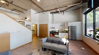 This striking urban loft showcases an industrial-modern aesthetic defined by dramatic double-height ceilings with exposed concrete slab, visible ductwork, and overhead conduit running throughout. The open-plan main level seamlessly connects the living area, dining space, and kitchen, creating a versatile and airy layout. Light hardwood flooring flows continuously underfoot, complementing the crisp white walls and raw architectural elements above.

The living area is generously proportioned, anchored by a contemporary sofa and lounge chair set atop a geometric area rug. A wall of oversized floor-to-ceiling windows and a glass entry door floods the space with abundant natural light while framing views of the surrounding streetscape and tree canopy. An exposed concrete column adds a sculptural industrial touch to the room.

The kitchen is efficiently designed with sleek white cabinetry extending to the ceiling, dark countertops, a tile backsplash, and a full suite of stainless steel appliances including a refrigerator, range, and dishwasher. A compact dining area with a table and colorful chairs sits adjacent.

A staircase with warm wood detailing leads to an upper loft level, visible from below, adding verticality and functional versatility to the space. The bathroom features a clean, modern vanity with a dark countertop, integrated sink, large mirror, and warm wood-tone flooring, conveying a calm and well-appointed finish throughout.