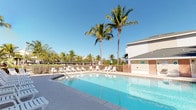 This outdoor pool facility offers a well-appointed recreational space designed for relaxation and leisure in a tropical setting. The centerpiece is a generously sized swimming pool with clear, blue water, reaching a depth of approximately 5 feet 6 inches at its deepest marked point. The pool features a gradual entry point, making it accessible for a range of swimmers. Surrounding the pool is a broad, open concrete sun deck that provides ample space for lounging and circulation. The deck is furnished with numerous white lounge chairs and upright seating, positioned along the poolside and adjacent to the main building to take full advantage of the sunny outdoor environment. Tall, mature palm trees are scattered throughout the area, lending a distinctly tropical ambiance while providing natural shade. A wood-paneled fence encloses the perimeter, ensuring privacy and safety. The adjacent single-story building features light-colored lap siding, a pitched roof, and large glass double doors that open directly onto the pool deck, seamlessly connecting indoor and outdoor spaces. Additional amenities, including restroom or utility facilities, are visible within the enclosure. The overall atmosphere is bright, open, and inviting, characteristic of a Florida-style resort or community recreational area.