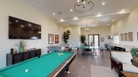 This single-floor recreation and social space is thoughtfully designed to accommodate a variety of leisure activities in a welcoming, well-appointed environment. The main game area is generously sized, anchored by a full-size pool table with a vibrant green felt surface and complemented by two foosball tables and a ping-pong table, making it a hub for friendly competition. Overhead, stylish pendant chandeliers and recessed lighting illuminate the space warmly, while tall ceilings contribute to an open, airy atmosphere. The lounge area offers plush, tufted leather sofas and accent chairs arranged around a rustic wood coffee table, creating an inviting retreat for conversation and relaxation. A gallery of framed patent blueprint prints adds a sophisticated, intellectual character to the walls. Tall indoor tropical plants are positioned throughout, adding organic texture and life to the space. A dedicated kitchenette along one wall features dark cabinetry, a smooth light-toned countertop, a stainless steel sink, built-in microwave, and floating display shelves — providing convenient refreshment options. French doors and additional entry doors connect the space to an outdoor area, allowing natural light to filter in and offering easy access to exterior amenities. The flooring transitions between wood-look tile and parquet-style hardwood, adding visual depth and warmth throughout.