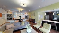 Located at 1019 Fox Hunt Ln in Raleigh, NC, this single-story home offers an open and inviting interior defined by warm hardwood flooring, soft yellow-green walls, and a cohesive color palette throughout. The main living area is spacious and thoughtfully arranged, centered around a plush gray sectional sofa and complemented by chartreuse accent chairs that add a touch of color and personality. A stylish drum flush-mount ceiling fixture anchors the room, while recessed lighting provides additional illumination across the open layout.

Built-in shelving with display niches lines one wall, offering both functional storage and decorative opportunity. A dark wood sideboard adds contrast against the lighter wall tones. The open floor plan connects the living area seamlessly to a dining space near a window-lined wall, allowing natural light to filter in and brighten the interior. The dining area benefits from views to the outdoors, enhancing the overall sense of openness.

A glimpse into the adjacent kitchen reveals pendant lighting and light countertops, suggesting a clean and functional design. The overall ambiance of the home is modern yet approachable, with carefully layered lighting and tasteful décor creating a warm, livable environment.