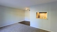 This single-floor residential space offers a clean, functional layout designed for comfortable everyday living. The main living area is generously proportioned, featuring plush wall-to-wall carpeting that transitions into a hard-surface flooring section, creating a natural visual distinction between living and dining zones. Neutral white walls throughout the space lend a bright, airy feel, while flush-mounted ceiling lighting provides consistent illumination. A standout architectural detail is the pass-through window connecting the living area to the kitchen, offering both a sense of openness and convenient accessibility between the two spaces. The kitchen, visible through the pass-through, is equipped with a refrigerator and includes countertop space, making it a practical and efficient area for meal preparation. Ceiling vents ensure proper climate control throughout the unit. The white baseboards and clean wall finishes contribute to a well-maintained, move-in-ready appearance. Overall, the space presents a straightforward and practical interior with a neutral palette that provides a versatile backdrop, easily adaptable to a variety of personal furnishing styles and décor preferences.