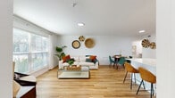 This well-appointed single-floor residence offers a cohesive blend of modern design and warm, bohemian-inspired decor. The open-concept living and dining area serves as the heart of the home, featuring warm-toned hardwood flooring that flows seamlessly throughout the space. Large windows allow abundant natural light to pour in, framing views of lush greenery outside. The living area is anchored by a comfortable neutral sofa accented with rust and teal cushions, a glass-top coffee table, and handwoven decorative wall pieces that add an artisanal touch.

Adjacent to the living space, the dining area accommodates a round wooden table surrounded by plush teal velvet chairs, creating an inviting setting for meals. The kitchen is equipped with stainless steel appliances, crisp white shaker cabinetry, a subway tile backsplash, and a clean white countertop with an undermount sink — all designed for both functionality and aesthetic appeal.

The bedroom features twin beds dressed in tropical-print bedding, complemented by rattan sunburst mirrors and warm wood furniture, creating a cheerful, relaxed atmosphere. Natural light filters through slatted blinds, adding to the airy feel. The two bathrooms are finished with marble-style tile surrounds — one featuring a glass-enclosed shower and the other a full soaking tub — both clean and elegantly appointed.