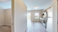 This single-floor unit offers a clean, contemporary layout designed for comfortable everyday living. Light wood-look flooring runs consistently throughout the space, creating a cohesive and airy aesthetic that complements the neutral warm-beige wall tones.

The kitchen is well-appointed with light wood cabinetry, granite-style countertops, a stainless steel refrigerator, electric range with over-the-range microwave, and a dishwasher. A window above the sink allows natural light to fill the workspace. The kitchen flows openly into the living area, making the combined space feel connected and functional.

The living area is generously sized, featuring a sliding glass door dressed with vertical blinds that leads to a private balcony, bringing in abundant natural light and outdoor views. A wall-mounted mini-split unit provides efficient climate control directly within the living space.

The bathroom continues the home's consistent design language with a single vanity topped with a granite-style countertop, an undermount sink, and light wood cabinetry with chrome hardware. A large mirror adds visual depth to the space.

A paneled interior door provides access to additional areas of the unit. Throughout the home, flush-mount ceiling fixtures provide clean, even lighting, and white baseboards offer a crisp finish to each room.