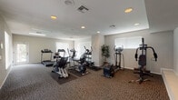 This fitness center offers a well-organized and comprehensively equipped workout environment spread across a single floor. The space is finished with durable, neutral-toned carpet throughout and features smooth white ceilings with recessed lighting that ensures consistent, even illumination across the entire floor plan. Multiple windows fitted with vertical blinds line the walls, allowing natural light to filter in and contribute to an open, airy atmosphere.

The cardio section is prominently featured and includes multiple treadmills, elliptical trainers, a stationary recumbent bike, and an exercise cycle, all arranged with ample spacing to allow for comfortable use. A dedicated strength training area houses a multi-station cable and weight machine alongside a rack of dumbbells, catering to a variety of resistance training routines. Rubber floor mats beneath the equipment provide protection and stability.

A glass-paneled exterior door provides direct access to the outdoors, where glimpses of a pool area can be seen, adding a refreshing connection to additional amenity spaces. The overall layout is practical and spacious, with clear circulation paths between equipment zones. Decorative potted plants add subtle warmth to the otherwise clean and functional aesthetic, making this fitness center a well-rounded facility for daily exercise needs.
