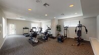 This fitness center offers a well-organized and comprehensively equipped workout environment spread across a single floor. The space is finished with durable, neutral-toned carpet throughout and features smooth white ceilings with recessed lighting that ensures consistent, even illumination across the entire floor plan. Multiple windows fitted with vertical blinds line the walls, allowing natural light to filter in and contribute to an open, airy atmosphere.

The cardio section is prominently featured and includes multiple treadmills, elliptical trainers, a stationary recumbent bike, and an exercise cycle, all arranged with ample spacing to allow for comfortable use. A dedicated strength training area houses a multi-station cable and weight machine alongside a rack of dumbbells, catering to a variety of resistance training routines. Rubber floor mats beneath the equipment provide protection and stability.

A glass-paneled exterior door provides direct access to the outdoors, where glimpses of a pool area can be seen, adding a refreshing connection to additional amenity spaces. The overall layout is practical and spacious, with clear circulation paths between equipment zones. Decorative potted plants add subtle warmth to the otherwise clean and functional aesthetic, making this fitness center a well-rounded facility for daily exercise needs.