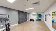 This purpose-built fitness studio offers a thoughtfully designed exercise space that maximizes functionality within a compact footprint. The room is defined by its standout "Freedom Climber" feature wall — a textured climbing panel embedded with colorful handholds arranged in a circular pattern, providing a unique and engaging upper-body workout option. The studio's interior is finished with a neutral color palette of whites and cool greys, complemented by herringbone-patterned light wood flooring that adds a modern, polished aesthetic throughout.

Recessed and panel ceiling lights provide bright, even illumination across the entire space. On one side, stationary cycling bikes with integrated monitor screens are positioned for cardio training. The opposite corner houses a well-organized equipment rack stocked with stability balls in multiple sizes and weighted medicine balls, alongside a flat storage bench for additional versatility. A wall-mounted mirror enhances the sense of space and supports form-checking during workouts.

Motivational wall graphics, including the bold "Challenge Your Limits" typography, reinforce the energetic atmosphere. Black-framed glass partition walls and a glazed door connect the studio to an adjacent, larger fitness area visible beyond, creating a sense of continuity. This studio is a well-appointed, multi-functional fitness environment suited for cycling, climbing, and functional training.
