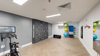 This purpose-built fitness studio offers a thoughtfully designed exercise space that maximizes functionality within a compact footprint. The room is defined by its standout "Freedom Climber" feature wall — a textured climbing panel embedded with colorful handholds arranged in a circular pattern, providing a unique and engaging upper-body workout option. The studio's interior is finished with a neutral color palette of whites and cool greys, complemented by herringbone-patterned light wood flooring that adds a modern, polished aesthetic throughout.

Recessed and panel ceiling lights provide bright, even illumination across the entire space. On one side, stationary cycling bikes with integrated monitor screens are positioned for cardio training. The opposite corner houses a well-organized equipment rack stocked with stability balls in multiple sizes and weighted medicine balls, alongside a flat storage bench for additional versatility. A wall-mounted mirror enhances the sense of space and supports form-checking during workouts.

Motivational wall graphics, including the bold "Challenge Your Limits" typography, reinforce the energetic atmosphere. Black-framed glass partition walls and a glazed door connect the studio to an adjacent, larger fitness area visible beyond, creating a sense of continuity. This studio is a well-appointed, multi-functional fitness environment suited for cycling, climbing, and functional training.