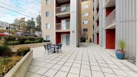 This ground-floor unit is distinguished by its expansive private patio, which serves as a central outdoor living area within a contemporary multi-story residential complex. The building's exterior showcases a modern design palette, combining warm beige horizontal siding with bold red accent panels that frame the unit's entry points and sliding glass doors, creating strong visual contrast throughout the courtyard-style setting.

The patio is finished with large-format concrete pavers laid in a clean grid pattern, providing a durable and visually cohesive surface across the generous outdoor area. A sliding glass door serves as the primary connection between the interior and the patio, allowing for easy indoor-outdoor access and natural light. The red-paneled exterior walls frame the entrance, adding a striking architectural element at ground level.

The outdoor space accommodates outdoor furniture, as evidenced by seating arrangements visible throughout, making it well-suited for relaxation and entertaining. Landscaping details include raised stone planters with decorative river rock, ornamental grasses, and small trees that add natural texture to the hardscaped environment. The patio is sheltered on multiple sides by the building's upper floors and balconies above, providing a sense of enclosure while remaining open to the sky and natural light.