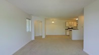 This single-floor space is designed with an open-concept layout that seamlessly connects the living area to the kitchen, creating a sense of spaciousness and ease of movement. The living area features plush, light-toned carpeting that extends throughout the space, contributing to a warm and comfortable atmosphere. Neutral white and beige walls provide a clean, versatile backdrop, while a textured ceiling adds a subtle architectural detail.

An arched passageway serves as an elegant architectural divider, leading toward what appears to be additional rooms or storage areas, adding both visual interest and functional separation within the open plan. A window on the far wall allows natural light to filter into the living space.

The kitchen is positioned at the rear of the open layout and is equipped with white cabinetry, a built-in microwave, an electric range, and dark countertops that provide a subtle contrast to the lighter elements. A pendant light fixture hangs above, adding a focused point of illumination. The kitchen also features a partial counter or bar area, offering a natural boundary between the cooking and living spaces while maintaining the overall open feel.