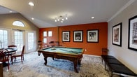 This single-floor space is designed as a dedicated entertainment and game room, centered around a solid wood pool table with ornate carved legs and a teal felt surface. The room's most striking visual element is its bold burnt-orange accent wall, which serves as a dramatic backdrop for a trio of framed black-and-white sports photographs, adding a gallery-like quality to the space. Vaulted ceilings contribute a sense of openness and volume, enhanced by a combination of recessed lighting and a multi-head track light fixture that ensures the playing surface is well illuminated. The flooring is covered with a large patterned area rug in muted blue and grey tones, providing a stylish contrast to the warm wall color. Natural light enters through several windows and a glass-panel door, brightening the space throughout the day. A bar-height counter with stools is positioned near the windows, offering a casual seating area. A side cabinet provides additional storage and surface space. Overall, the room balances a sporty, social atmosphere with thoughtful design details.