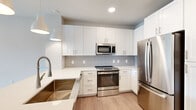 This single-story unit at Lenox in Houston, Texas offers a clean, modern design with thoughtfully selected finishes throughout. The kitchen serves as a central highlight, outfitted with crisp white shaker-style cabinetry extending to the ceiling for ample storage, a soft gray subway tile backsplash, and a full suite of stainless steel appliances including a gas range, over-the-range microwave, and French door refrigerator. A deep undermount sink is set into white quartz countertops, while stylish pendant lights hang overhead, adding a refined touch. Light wood-look flooring runs through the main living areas, contributing to the unit's bright, contemporary feel.

The living area is open and versatile, with neutral walls and natural light entering through a large black-framed picture window that provides views of the surrounding streetscape. A ceiling fan is installed overhead for added comfort. The bedroom features plush carpet flooring, a generous closet with a paneled door, and its own large black-framed window, maintaining the consistent modern aesthetic. The unit's overall palette of whites, soft grays, and warm wood tones creates a cohesive, welcoming atmosphere throughout.