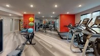 This fitness center is designed to accommodate a wide range of workout styles and fitness goals within a single, open-plan space. The room is finished with light wood-look flooring that lends a clean, contemporary aesthetic throughout. Recessed lighting illuminates the entire area evenly, while large windows along one wall allow natural light to fill the space and provide views to the exterior.

The cardio zone features a lineup of treadmills, elliptical trainers, stationary bikes, and rowing machines — including Matrix-branded equipment — offering extensive options for aerobic training. Large flat-screen TVs are mounted on the walls, keeping members entertained during their workouts. Floor-to-ceiling mirrors on multiple walls enhance the sense of openness while allowing users to monitor their form.

The strength and functional training area is equally well-appointed, with cable machines, a leg press, a multi-station weight system, and a full rack of dumbbells. Bold red accent walls energize this section of the space, adding visual contrast to the otherwise neutral color palette. A dedicated functional fitness zone includes stability balls in multiple sizes, resistance accessories, and foam rollers neatly organized on a wall-mounted rack. The overall layout is thoughtfully arranged to ensure ample room for movement between stations, creating an accessible and motivating environment for all fitness levels.