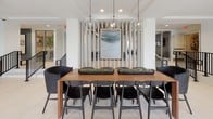 This single-floor space is designed with a clean, contemporary aesthetic that prioritizes openness and natural light. The lobby area serves as a welcoming entry point, featuring large floor-to-ceiling windows framed in dark metal that flood the interior with daylight and offer views of mature trees outside. Large-format white tile flooring extends throughout, lending a cohesive and polished look across all areas.

The seating area near the entrance is furnished with a low-profile gray sofa, a warm terracotta accent chair, and a solid wood coffee table, creating a comfortable and inviting gathering spot. A glass vestibule at the main entrance provides a secure and elegant transition into the space.

A built-in coffee and refreshment bar is thoughtfully integrated into one section of the floor, featuring wood cabinetry with matte black hardware, open white shelving, a wall-mounted display screen, and a dark tile backsplash that adds texture and contrast.

Adjacent to this is a communal dining area anchored by a substantial wood table surrounded by black woven chairs. Globe pendant lights hang overhead, and a vertical slatted screen with framed artwork serves as a refined architectural divider. The overall ambiance is bright, modern, and functional, designed to foster both casual interaction and focused use.