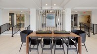 This single-floor space is designed with a clean, contemporary aesthetic that prioritizes openness and natural light. The lobby area serves as a welcoming entry point, featuring large floor-to-ceiling windows framed in dark metal that flood the interior with daylight and offer views of mature trees outside. Large-format white tile flooring extends throughout, lending a cohesive and polished look across all areas.

The seating area near the entrance is furnished with a low-profile gray sofa, a warm terracotta accent chair, and a solid wood coffee table, creating a comfortable and inviting gathering spot. A glass vestibule at the main entrance provides a secure and elegant transition into the space.

A built-in coffee and refreshment bar is thoughtfully integrated into one section of the floor, featuring wood cabinetry with matte black hardware, open white shelving, a wall-mounted display screen, and a dark tile backsplash that adds texture and contrast.

Adjacent to this is a communal dining area anchored by a substantial wood table surrounded by black woven chairs. Globe pendant lights hang overhead, and a vertical slatted screen with framed artwork serves as a refined architectural divider. The overall ambiance is bright, modern, and functional, designed to foster both casual interaction and focused use.