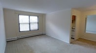 This single-floor residential unit offers a clean, functional layout designed for comfortable everyday living. The main living area is finished with wall-to-wall carpeting in a neutral tone, providing a warm and quiet underfoot experience. A large window along one wall allows natural light to filter in, offering views of the surrounding urban cityscape. A baseboard heating system runs along the perimeter of the room, ensuring consistent warmth throughout the space. The walls are finished in a crisp white, creating a bright and open atmosphere that allows for versatile interior styling. Transitioning from the living area, the adjoining kitchen is accessible through a wide opening, making the layout feel connected and practical. The kitchen features wood-toned upper cabinetry and includes a freestanding gas range with an oven, providing a functional cooking space. The kitchen flooring transitions to a hard surface, distinguishing it from the carpeted living area. The overall design of the unit is straightforward and utilitarian, with clean lines and a neutral palette throughout, making it easy to personalize. The presence of urban views through the windows speaks to the building's elevated positioning within its city environment.