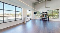 Located at 6050 Roswell Rd in Sandy Springs, Georgia, this contemporary fitness and dance studio offers a thoughtfully designed, open-plan space ideal for a wide range of movement-based activities. The single-floor layout maximizes usable floor space, with smooth hardwood flooring that extends throughout the entire area, providing an ideal surface for exercise, dance, and training.

A defining feature of the studio is the expansive floor-to-ceiling mirror wall, which spans the full width of one side of the space, enhancing the sense of openness and making the room feel even more generous in scale. Large, black-framed grid windows line the walls on multiple sides, flooding the interior with natural light and offering views of the surrounding outdoor environment.

The ceiling is notably high, with an exposed industrial-style design that incorporates linear LED lighting fixtures and ceiling fans, contributing both to the modern aesthetic and practical functionality of the space. The crisp white walls and clean architectural lines give the studio a bright, professional atmosphere.

A dedicated fitness area is equipped with a wall-mounted television, medicine balls, stability balls, kettlebells, and additional training equipment stored on a rack. A secondary open zone provides ample room for group classes, yoga, dance, or personal training sessions. The overall ambiance is clean, energetic, and professional — well-suited for a dynamic fitness environment.