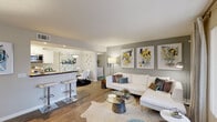 This thoughtfully designed single-floor residence offers a seamless blend of modern style and functional living. The open-concept living area is anchored by rich hardwood-style flooring, neutral gray accent walls, and large windows that invite natural light throughout. A white sectional sofa, glass coffee table, and a bold triptych of abstract artwork create a refined yet welcoming atmosphere. The living space flows effortlessly into a bar-height kitchen pass-through and dining area, ideal for entertaining.

The kitchen features white cabinetry with dark granite-style countertops, stainless steel appliances including a gas range and built-in microwave, and light wood-look flooring for a clean, contemporary finish.

The bedrooms are standout spaces: one features a dramatic navy blue textured accent wall flanked by floor-length mirrors, a ceiling fan, and plush carpet; the other showcases a full-wall ocean-inspired mural in soft blue and earth tones, filling the room with a serene, artistic presence. A walk-in closet with wire shelving provides generous storage, conveniently adjacent to a full bathroom.

The bathroom is finished with wood-look tile flooring, a floating vanity with dark countertop, an undermount sink, and a shower-tub combination, completing this well-appointed home.
