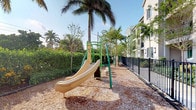 This outdoor playground area offers a well-appointed recreational space designed for children, situated within a lushly landscaped setting. The play structure, finished in teal and cream tones, features a wide slide and climbing elements, all set atop a generous wood chip surface that provides both safety and a natural aesthetic. Tall, swaying palm trees frame the space overhead, lending a distinctly tropical character to the environment. Dense hedgerows and mature tropical foliage line the perimeter, creating a sense of enclosure and privacy from the surrounding area. A sleek black iron fence runs along one side of the playground, defining the boundary between the recreational space and the adjacent multi-story building. Additional play elements are visible further along the pathway, suggesting a thoughtfully extended play zone. The overall ambiance is bright and inviting, benefiting from ample natural sunlight filtering through the palm canopy. The combination of verdant plantings, durable equipment, and safety-conscious ground cover makes this an attractive and functional outdoor space well-suited for active, outdoor play in a warm, tropical setting.