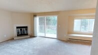 Located at 9823 NE 130th Pl, Unit 3205 in Kirkland, Washington, this single-floor residence offers a comfortable and well-lit living environment surrounded by lush Pacific Northwest greenery. The spacious living room serves as the heart of the home, finished with plush wall-to-wall carpeting and neutral-toned walls that create a warm, inviting atmosphere. A tile-surround fireplace anchors one side of the room, providing both a functional heating source and an elegant focal point. Natural light pours generously into the space through a large sliding glass door that opens directly onto a private balcony, where views of mature trees and green landscaping create a serene outdoor connection. A broad picture window on the adjacent wall further enhances the brightness of the room and features a built-in window seat — a charming architectural detail ideal for relaxing or additional seating. Baseboard heating runs beneath the windows, ensuring year-round comfort. The textured ceiling adds a classic finishing touch throughout. The overall ambiance is bright, open, and tranquil, making this Kirkland residence a peaceful retreat with practical features and a strong connection to the natural surroundings.
