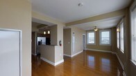 This single-floor space offers an open, airy layout designed for comfortable everyday living. The living area is anchored by rich, polished hardwood flooring that reflects natural light streaming in through large, double-hung windows dressed with horizontal blinds. A ceiling fan overhead adds both function and a classic finishing touch to the main living space.

The kitchen is partially separated from the living area by a raised breakfast bar counter, creating a natural division while maintaining an open feel. Dark-stained cabinetry lines the kitchen walls, complemented by a stainless steel refrigerator and pendant lighting that hangs above the counter, adding warmth to the cooking space.

Warm, earthy tones define the walls throughout, giving the interior a cohesive and welcoming ambiance. Structural columns and a wide archway frame the transition between the kitchen and living areas, adding architectural character without compromising the sense of openness.

A doorway and additional entry points visible along the back wall suggest further rooms beyond the main living space. Clean white trim and baseboards provide a crisp contrast to the warm wall tones, while practical details such as a fire extinguisher and utility panels reflect a thoughtfully maintained environment. Overall, the space offers a well-balanced blend of functionality and comfortable, modern design.
