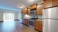 Located at 1699 Stieger Lake Ln in Victoria, MN, this two-bedroom, one-bathroom residence offers a well-designed single-floor layout with a thoughtful mix of finishes and functional spaces. The kitchen is a standout feature, showcasing warm honey-toned cabinetry with built-in wine rack detail, granite countertops, and a full suite of stainless steel appliances including a gas range, built-in microwave, dishwasher, and refrigerator. Rich hardwood-style flooring flows throughout the kitchen and dining area, creating a cohesive and inviting atmosphere. The living and dining area benefits from natural light streaming through a large sliding glass door fitted with vertical blinds, offering a pleasant outdoor view of surrounding trees. The primary bedroom is a comfortable, well-proportioned room with soft carpeting and dual windows that bring in additional natural light. The second bedroom also features hardwood-style flooring and access to a sliding glass door. The bathroom is finished with matching granite countertops, a warm wood vanity, a large mirror, and hardwood-style flooring. A dedicated in-unit laundry room includes a full-size washer and dryer with wire shelving for added storage. Dark wood baseboards throughout the unit add a polished, finishing touch to every room.