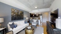 This contemporary single-floor residence showcases a thoughtfully designed open-concept layout that effortlessly connects the living area and kitchen into one cohesive, flowing space. The most striking architectural feature is the exposed concrete ceiling, which lends an industrial character and urban sophistication to the interior. Warm hardwood flooring runs throughout, providing a rich contrast to the cooler tones above.

The living area is anchored by a plush white sofa, complemented by elegant accent chairs and a distinctive gold geometric coffee table. A large, dramatic abstract artwork in deep blues and golds dominates a slate-gray feature wall, adding a bold artistic focal point. Soft, layered lighting enhances the ambiance throughout.

The kitchen is open and functional, featuring dark cabinetry, granite countertops, and stainless steel appliances. A breakfast bar with seating offers a casual dining option while maintaining the open sightlines of the space. Pendant lighting above the bar adds warmth and definition to the kitchen zone.

Overall, this space strikes a compelling balance between industrial design and refined, modern furnishings, creating an inviting and visually dynamic environment well-suited for both everyday living and entertaining.