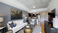 This contemporary single-floor residence showcases a thoughtfully designed open-concept layout that effortlessly connects the living area and kitchen into one cohesive, flowing space. The most striking architectural feature is the exposed concrete ceiling, which lends an industrial character and urban sophistication to the interior. Warm hardwood flooring runs throughout, providing a rich contrast to the cooler tones above.

The living area is anchored by a plush white sofa, complemented by elegant accent chairs and a distinctive gold geometric coffee table. A large, dramatic abstract artwork in deep blues and golds dominates a slate-gray feature wall, adding a bold artistic focal point. Soft, layered lighting enhances the ambiance throughout.

The kitchen is open and functional, featuring dark cabinetry, granite countertops, and stainless steel appliances. A breakfast bar with seating offers a casual dining option while maintaining the open sightlines of the space. Pendant lighting above the bar adds warmth and definition to the kitchen zone.

Overall, this space strikes a compelling balance between industrial design and refined, modern furnishings, creating an inviting and visually dynamic environment well-suited for both everyday living and entertaining.