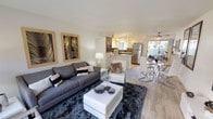 This well-appointed single-floor residence offers an open-concept layout that seamlessly connects the living, dining, and kitchen areas into one cohesive and functional space. The living area is anchored by a plush gray sofa and accented with a dark shag area rug, a sleek white coffee table, and contemporary botanical artwork in warm wood frames that add a natural touch to the neutral palette. Light wood-style flooring runs throughout, lending warmth and continuity to the space. The kitchen features stainless steel appliances, white cabinetry, a mosaic tile backsplash, and a breakfast bar that provides additional seating and workspace. Track lighting above ensures the kitchen remains bright and practical. The dining area is positioned near expansive sliding glass doors that flood the space with natural light and open to an outdoor area, creating an airy, open ambiance. A ceiling fan overhead adds comfort to the dining zone. The overall design aesthetic is modern and clean, with a carefully curated mix of textures and neutral tones that create an inviting yet sophisticated atmosphere throughout the entire living space.
