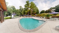 Located at 3000 Shadowood Pkwy SE in Atlanta, Georgia, this outdoor pool area offers a well-appointed recreational retreat set against a backdrop of mature trees and manicured landscaping. The centerpiece of the space is a generously sized freeform swimming pool with clear, turquoise water, complemented by a separate, smaller wading or spa pool that features a decorative tiled border. The pool deck is finished in neutral concrete pavers, providing ample space for lounging throughout the day. Numerous chaise lounge chairs are arranged around the perimeter of the pool, offering comfortable sunbathing options, while multiple purple patio umbrellas provide shaded seating areas with accompanying tables and chairs for those seeking relief from the sun. A covered pavilion with a wood-framed, pitched roof anchors one side of the deck, offering additional shade and a defined gathering space. The surrounding landscape features tiered garden beds filled with flowering plants, ornamental grasses, and dense greenery, creating a natural, park-like atmosphere. Tall, established trees frame the entire area, enhancing privacy and contributing to the tranquil ambiance of this outdoor amenity space.