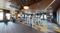 This expansive, single-level fitness center delivers a visually dynamic workout environment that blends modern amenities with bold, retro-inspired design. The space is divided into distinct functional zones, each thoughtfully appointed for a comprehensive fitness experience.

The main cardio and strength area features a generous array of Life Fitness treadmills, elliptical trainers, and cable machines, arranged across a geometric patterned carpet in vivid yellow, grey, and taupe tones. Wood-paneled ceilings with angular LED linear lighting illuminate this zone, lending warmth and a polished, contemporary feel. Full-length mirrors along the walls amplify the sense of openness and allow users to monitor their form throughout workouts.

A dedicated free weights section includes a full dumbbell rack and adjustable benches, positioned alongside floor-to-ceiling windows that invite natural light and outdoor views. Adjacent to this is a functional training zone with dark hardwood flooring, featuring TRX suspension systems, kettlebells, medicine balls, and stability equipment neatly organized on wall-mounted racks. A wall-mounted flat-screen display provides instructional content in this area.

The standout design element throughout is the vivid turquoise ceiling adorned with vintage signage artwork — including references to classic neon signs — accented by large ceiling fans and globe pendant lights. This artistic ceiling treatment creates a lively, energetic atmosphere that sets this fitness center apart.