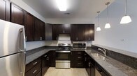 This single-floor residence offers a thoughtfully designed layout that balances comfort and functionality. The kitchen stands out as a focal point of the home, showcasing rich dark espresso cabinetry that extends to the ceiling, providing generous storage throughout. Granite countertops in a speckled dark tone offer ample workspace, while stainless steel appliances — including a gas range, built-in microwave, and undermount sink — lend a sleek, contemporary finish. Light wood-style flooring in the kitchen enhances the contrast against the dark cabinetry, and pendant lighting adds a refined touch above the counter area. A hallway connects the living spaces seamlessly, with neutral warm-toned walls and clean white trim creating a cohesive and inviting atmosphere throughout. The living area features plush wall-to-wall carpeting underfoot and benefits from a ceiling fan that promotes year-round comfort. A broad window fills the room with natural light, brightening the space and creating an open, airy feel. The overall design aesthetic is modern and clean, with consistent finishes that give the home a polished, move-in-ready appeal.