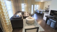 This single-floor space showcases a thoughtfully designed open-concept layout that connects the living and dining areas in a seamless, functional flow. Rich, dark hardwood flooring runs throughout, grounding the space with warmth and a sense of continuity.

The living area is anchored by a generously sized dark sectional sofa accented with layered throw pillows and a plaid blanket, centered around a distinctive round upholstered ottoman that doubles as a coffee table. A bold geometric-patterned area rug defines the seating zone, while a sleek arc floor lamp provides ambient lighting overhead. Large windows dressed in striking gold and white chevron-patterned curtains allow natural light to fill the room and contribute to the space's lively, contemporary aesthetic.

The dining area, visible from the living room, features a round dining table surrounded by upholstered chairs in a neutral tone, complemented by a simple floral arrangement as a centerpiece. The open sightlines between both areas enhance the sense of spaciousness throughout.

The overall ambiance is modern and inviting, with a carefully curated mix of bold patterns, neutral base tones, and warm wood finishes that come together to create a cohesive and stylish interior environment.