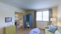 This single-floor space offers a practical and lightly decorated layout with a warm, casual ambiance. The main living area is finished with wall-to-wall carpeting and painted in neutral tones, creating a clean and understated backdrop. A decorative blue folding screen serves as a stylish room divider, adding a touch of character to the space. A wall-mounted air conditioning unit and baseboard heating provide year-round climate control. Natural light enters through a generously sized window dressed with floor-length curtains, brightening the room. The living area is furnished with a rounded upholstered ottoman and a delicate wrought-iron accent chair, contributing to the eclectic aesthetic. An open doorway leads directly into the kitchen, which features light wood cabinetry, a white stove, and a compact, functional layout. A small dining area with a table and chairs is visible adjacent to the kitchen, making the transition between spaces seamless. A yellow chest of drawers near the entry adds a pop of color. Overall, the space presents a cozy and functional single-level layout suited for everyday living.