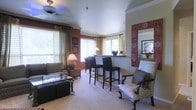 This single-story space offers an open-concept layout that seamlessly connects the living area to a raised breakfast bar. The living room is furnished with a cushioned sofa, an ornate accent chair with carved wooden legs, and a sleek glass-top coffee table, creating a balanced mix of traditional and contemporary styles. Crown molding frames the smooth white ceiling, which is fitted with a ceiling fan, adding both function and a classic finishing touch.

Large windows dressed with layered curtains and horizontal blinds allow natural light to fill the space while maintaining privacy. The warm, earthy wall tones — including a rich terracotta accent wall — add depth and character. A striking decorative mirror with an ornate red frame serves as a bold focal point, complementing the overall warm color palette.

The breakfast bar area features tall bar stools with dark upholstered seats and a pendant light overhead, making it an ideal casual dining or entertaining spot. A table lamp in the corner adds warm ambient lighting. Carpeted flooring runs throughout, contributing to a cozy and inviting atmosphere overall.