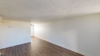 Located at 1220 E Medlock Dr in Phoenix, AZ, this single-story unit offers a straightforward and functional layout designed for comfortable everyday living. The open living area is finished with rich, dark wood-style flooring that extends throughout the main spaces, complemented by crisp white walls and clean baseboard trim that give the interior a bright, neutral aesthetic. Large vertical blinds in the living room allow for natural light control while framing a glass-paneled exterior door that connects the space to the outside.

The bedrooms are carpeted and feature vertical blinds on the windows, with one room equipped with a ceiling fan and light fixture for added comfort. The rooms offer a clean, open feel with ample wall space for flexible furniture arrangements.

A compact hallway connects the main living areas to the bathroom and entry, where a wall-mounted sconce provides warm ambient lighting. The hallway also features a wall panel unit, likely for HVAC or utility access. The full bathroom includes a tub/shower combo with white tile surround, a toilet, and a vanity, all finished with wood-style flooring matching the rest of the unit. The kitchen, visible in the dollhouse view, is efficiently positioned adjacent to the living space, completing this well-organized single-floor layout.