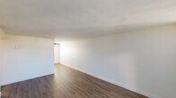 Located at 1220 E Medlock Dr in Phoenix, AZ, this single-story unit offers a straightforward and functional layout designed for comfortable everyday living. The open living area is finished with rich, dark wood-style flooring that extends throughout the main spaces, complemented by crisp white walls and clean baseboard trim that give the interior a bright, neutral aesthetic. Large vertical blinds in the living room allow for natural light control while framing a glass-paneled exterior door that connects the space to the outside.

The bedrooms are carpeted and feature vertical blinds on the windows, with one room equipped with a ceiling fan and light fixture for added comfort. The rooms offer a clean, open feel with ample wall space for flexible furniture arrangements.

A compact hallway connects the main living areas to the bathroom and entry, where a wall-mounted sconce provides warm ambient lighting. The hallway also features a wall panel unit, likely for HVAC or utility access. The full bathroom includes a tub/shower combo with white tile surround, a toilet, and a vanity, all finished with wood-style flooring matching the rest of the unit. The kitchen, visible in the dollhouse view, is efficiently positioned adjacent to the living space, completing this well-organized single-floor layout.