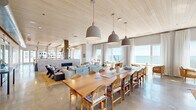 This single-floor coastal retreat is designed to impress, combining sophisticated beachside aesthetics with generous, open-plan living. Whitewashed wood-paneled ceilings run throughout, lending warmth and a natural, airy character to the entire space. Polished concrete floors provide a sleek, modern foundation that complements the organic materials and coastal palette.

The expansive main hall centers around a long communal dining table surrounded by woven rattan chairs, illuminated by a row of oversized grey dome pendant lights. Floor-to-ceiling windows and sheer linen drapes frame breathtaking panoramic views of the ocean, flooding the interior with natural light. A seating lounge area anchors one end of the hall, featuring a sleek linear stone fireplace with a striking dark metal surround, flanked by curated furnishings in neutral and earthy tones.

A dedicated bar and kitchen area is outfitted with stone countertops, stainless steel appliances, a draft beer system, and exposed beam detailing with warm string lighting overhead. A secondary lounge space opens directly onto an outdoor terrace via large glass-paned doors, offering access to an exterior entertaining area with water views. Throughout, colorful original artwork punctuates the whitened wood walls, adding a vibrant, creative energy to this refined coastal venue.
