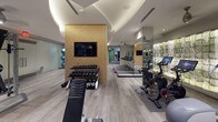 Located at 2009 N 14th St in Arlington, VA, this well-appointed fitness center offers a comprehensive workout environment designed with both functionality and aesthetics in mind. The space is organized across a single floor and divided into distinct training zones, each thoughtfully equipped for a variety of fitness activities.

The main cardio and strength area features light wood-look flooring, recessed lighting, and a striking curved ceiling detail that adds architectural character. A row of Precor treadmills lines one wall, backed by a textured, padded accent wall that doubles as a design focal point. Elliptical machines, cable weight systems, adjustable benches, and a full dumbbell rack round out the strength and conditioning offerings. A wall-mounted flat-screen TV is embedded into a natural cork accent wall, adding warmth to the space.

A dedicated boxing and functional training zone features rubberized flooring, a freestanding heavy bag and speed bag station, and floor-to-ceiling mirrors that amplify the sense of space. Peloton bikes provide additional cardio options nearby.

Glass-paneled doors with motivational text — "Sweat. Smile. Repeat." — separate zones while maintaining visual openness. Strategic mirror placement throughout enhances the spacious feel, and ceiling fans complement the recessed lighting for a comfortable training atmosphere. The overall design balances modern finishes with a practical, performance-focused layout.