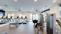 Located in Orlando, FL, this modern fitness center offers a comprehensive workout facility designed to support a full range of exercise routines. The space is spread across a single floor and is organized into distinct zones, accommodating both cardio and strength training needs. The cardio area features multiple Matrix treadmills and stair climbers, positioned near large windows and glass doors that invite ample natural light and provide views of the surrounding outdoor greenery and pool area. Ceiling fans are mounted throughout, enhancing airflow and comfort during workouts. The strength training zone is well-appointed with a variety of Matrix cable and selectorized machines, a multi-station rack with free weights, dumbbells on tiered racks, and adjustable benches. Durable rubberized flooring covers the entire space, offering both safety and comfort underfoot. A striking large-scale black-and-white photographic mural decorates one wall, adding a dynamic and artistic focal point to the space. Recessed lighting combined with natural light creates a bright, energizing atmosphere throughout. Mirrors line select walls, enhancing the sense of openness and allowing exercisers to monitor their form. The overall design balances functionality with contemporary aesthetics, making this fitness center a standout amenity.