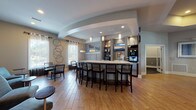 Located in Summerville, SC, this single-story community clubhouse at 10825 Dorchester Rd offers a well-appointed gathering and wellness facility. The centerpiece is a grand lounge area distinguished by soaring ceilings with exposed dark wood beam accents, an elegant chandelier, and a dramatic arched window wall that floods the space with natural light. Wood-look tile flooring flows throughout, complementing the sophisticated neutral color palette. A stone fireplace with a mounted TV anchors one end of the lounge, while plush seating arrangements create multiple conversation areas.

Adjacent to the lounge is a café-style bar and dining area featuring a long, light-surfaced counter with bar seating, pendant lighting, a mounted TV, and built-in shelving — ideal for casual gatherings. The space also includes a workstation area for residents' use.

The fitness wing is comprehensive, housing a main exercise room equipped with strength machines, ellipticals, stationary bikes, and a free-weight rack, all beneath high ceilings with ceiling fans and ample natural light from surrounding windows. A dedicated spin studio provides stationary cycling bikes, a wall-mounted TV, and a ceiling fan. A separate stretching and functional fitness studio features rubber flooring, medicine ball storage, exercise mats, a tray ceiling, and bright windows overlooking the outdoor pool area.