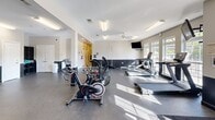 Located at 517 Yosef Dr in Boone, NC, this fitness center offers a generously sized, single-floor exercise space designed to accommodate a wide range of workout needs. The open floor plan provides ample room to move between equipment zones, with durable rubber flooring throughout that ensures both safety and comfort during exercise.

The cardio section is well-appointed with treadmills, stationary bikes, and elliptical-style machines positioned near large windows that allow natural light to fill the space. A ceiling fan overhead adds to the comfort of high-intensity workouts in this area.

The strength training section is equally comprehensive, featuring a cable and pulley multi-station machine, a power rack with barbells and weight plates, a full rack of dumbbells, and multiple selectorized resistance machines for targeted muscle training. Flat and adjustable benches are available throughout the space for added versatility.

Wall-mounted televisions are strategically placed to keep users entertained during their workouts. The interior is finished with a clean, neutral color palette — white upper walls accented by a dark lower wainscoting — giving the facility a modern and professional appearance. Fluorescent lighting ensures consistent illumination throughout the entire space, complementing the natural light from the windows. Restroom access is available within the facility.
