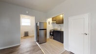 Located at 1093 Sheridan Ave in Elizabeth, NJ, this single-floor unit showcases a thoughtful renovation with clean, modern finishes throughout. Light gray luxury vinyl plank flooring runs continuously across the living areas, complementing the neutral wall tones and white trim work that give the space a fresh, cohesive look. The entry area greets you with a classic white door, a large window fitted with blinds, a cast-iron radiator, and a wrought-iron stair railing — a nod to the building's character. The spacious living area flows openly into an adjacent room, also fitted with a radiator and dual windows that allow generous natural light to pour in. The compact kitchen is efficiently designed, featuring dark shaker-style cabinetry, granite countertops, a stainless steel gas range, an undermount sink with a modern pull-down faucet, and a classic white subway tile backsplash. A full-size stainless steel refrigerator is conveniently positioned just outside the kitchen. The bathroom has been fully updated with large-format marble-look wall tiles surrounding the soaking tub and shower, light wood-look floor tiles, a dark vanity with a white countertop, and a wall-mounted mirror, delivering a clean and polished aesthetic throughout.