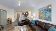 This single-floor residence showcases an open-concept layout that integrates the living area and kitchen into one cohesive, flowing space. The living area is anchored by a dark leather sofa and matching armchair with ottoman, arranged over a richly patterned area rug atop polished concrete floors. A rustic wood coffee table and vibrant abstract wall art add character and visual interest to the space. Track lighting along the ceiling provides adjustable illumination throughout the main living area. The kitchen features dark cabinetry, stainless steel appliances including a full-size refrigerator, and a peninsula with a stone-look countertop and bar seating for two. Pendant lights hang above the peninsula, adding warmth and definition to the kitchen zone. The neutral white walls and smooth ceilings create a clean backdrop that highlights the contemporary furnishings and décor. A hallway visible from the main area leads to additional rooms, with a wood-paneled door providing a subtle design accent. The overall ambiance is modern and urban, with a comfortable yet stylish aesthetic that balances function and design.