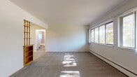 This 1-bedroom, 1-bathroom unit is laid out entirely on a single floor, offering a straightforward and efficient floor plan. The living area is spacious and carpeted, featuring a bank of large windows that allow ample natural light to fill the room and offer pleasant views of surrounding greenery. A baseboard heating system runs along the perimeter walls, and a wall-mounted air conditioning unit provides additional climate control. The entryway opens directly into this bright main living space.

The kitchen is efficiently designed with warm oak cabinetry providing generous storage above and below the counters. It is equipped with a gas range, a stainless steel sink, and laminate flooring, offering a practical and functional cooking space.

The bedroom is a comfortable, carpeted room with its own window that brings in natural light and outdoor views. A ceiling fan adds to the room's comfort and ventilation.

The bathroom features a full tiled tub-shower combination with light-colored wall tile, a toilet, and a vanity with mirror. A frosted window provides natural light while maintaining privacy. The floor is finished with a wood-look vinyl tile, completing the clean and functional design of this well-organized unit.
