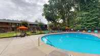 Located at 306 Estes Drive Ext in Carrboro, North Carolina, this outdoor pool area offers a well-appointed recreational retreat set against a backdrop of mature trees and natural greenery. The centerpiece of the space is a generously sized oval swimming pool with vivid blue water, trimmed with classic blue tile along its edges and equipped with stainless steel entry rails for easy access. The pool accommodates varying depths, as indicated by depth markers along its perimeter.

Surrounding the pool is a spacious concrete sun deck that provides ample room for relaxation and movement. Teal lounge chairs are arranged thoughtfully around the pool's edge, offering comfortable spots for sunbathing. Vibrant orange patio umbrellas paired with small bistro-style tables are positioned throughout the deck, providing shaded seating areas ideal for casual gatherings.

A brick accessory building anchors one side of the pool area, contributing a classic architectural character to the space. The entire area is enclosed by a black iron fence, ensuring safety and a defined boundary. Tall, leafy trees form a dense natural canopy along the perimeter, creating a sense of privacy and a cool, shaded atmosphere. The lush wooded surroundings enhance the tranquil, resort-like ambiance of this community outdoor space.