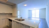 This single-floor space showcases a thoughtfully designed open-concept layout that seamlessly connects the kitchen and living areas. The kitchen is a standout feature, appointed with sleek granite countertops, a double stainless steel undermount sink, and contemporary cabinetry finished in a light wood tone with minimalist hardware. A matching granite backsplash adds a refined touch, while stainless steel appliances contribute to the modern aesthetic. The kitchen island flows naturally into the open living area, creating an ideal space for both everyday living and entertaining. The living area is bathed in natural light, courtesy of expansive floor-to-ceiling windows that span an entire wall, offering an open and airy atmosphere with views to an outdoor balcony. The clean white walls and smooth ceilings, fitted with recessed and flush-mount lighting, enhance the sense of brightness throughout. Neutral-toned flooring ties the spaces together cohesively, reinforcing the unit's contemporary and polished design. Overall, this space delivers a bright, functional, and modern living environment with a strong emphasis on open space and natural light.